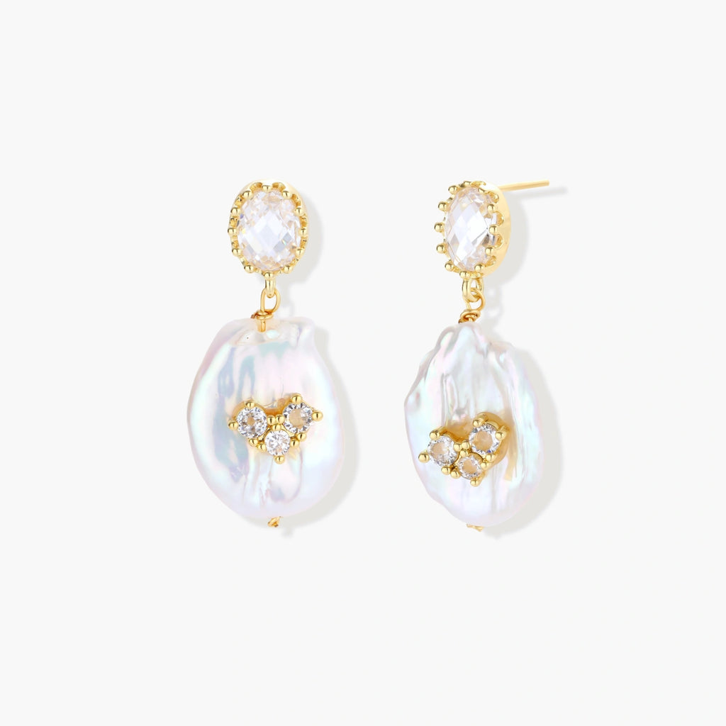 Gold-plated drop earrings with iridescent baroque pearls and crystal flower accents on cream background