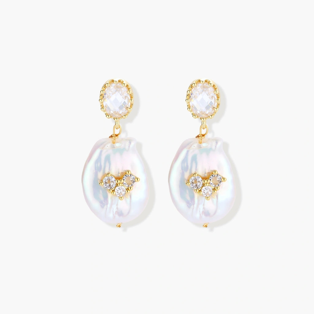 Gold-plated drop earrings with iridescent baroque pearls and crystal flower accents on cream background