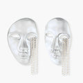 Silver face-shaped statement earrings with flowing chain tassels hanging from sculpted lips on cream background
